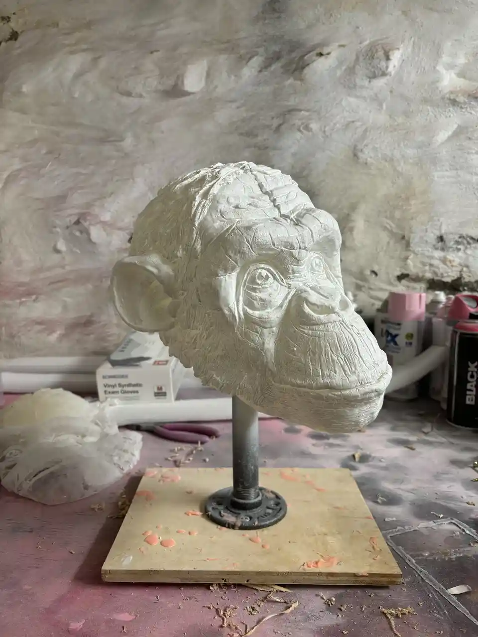 A white clay sculpture of a monkey's head on a metal stand sits on a messy art table with spray paint cans and gloves…