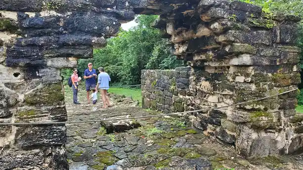A family of four stands inside ancient stone ruins with moss-covered walls and rope barriers, looking out toward lush…