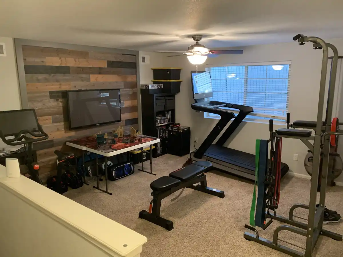 A well-equipped home gym in a basement features a treadmill, weight bench, squat rack, exercise bike, and resistance bands…