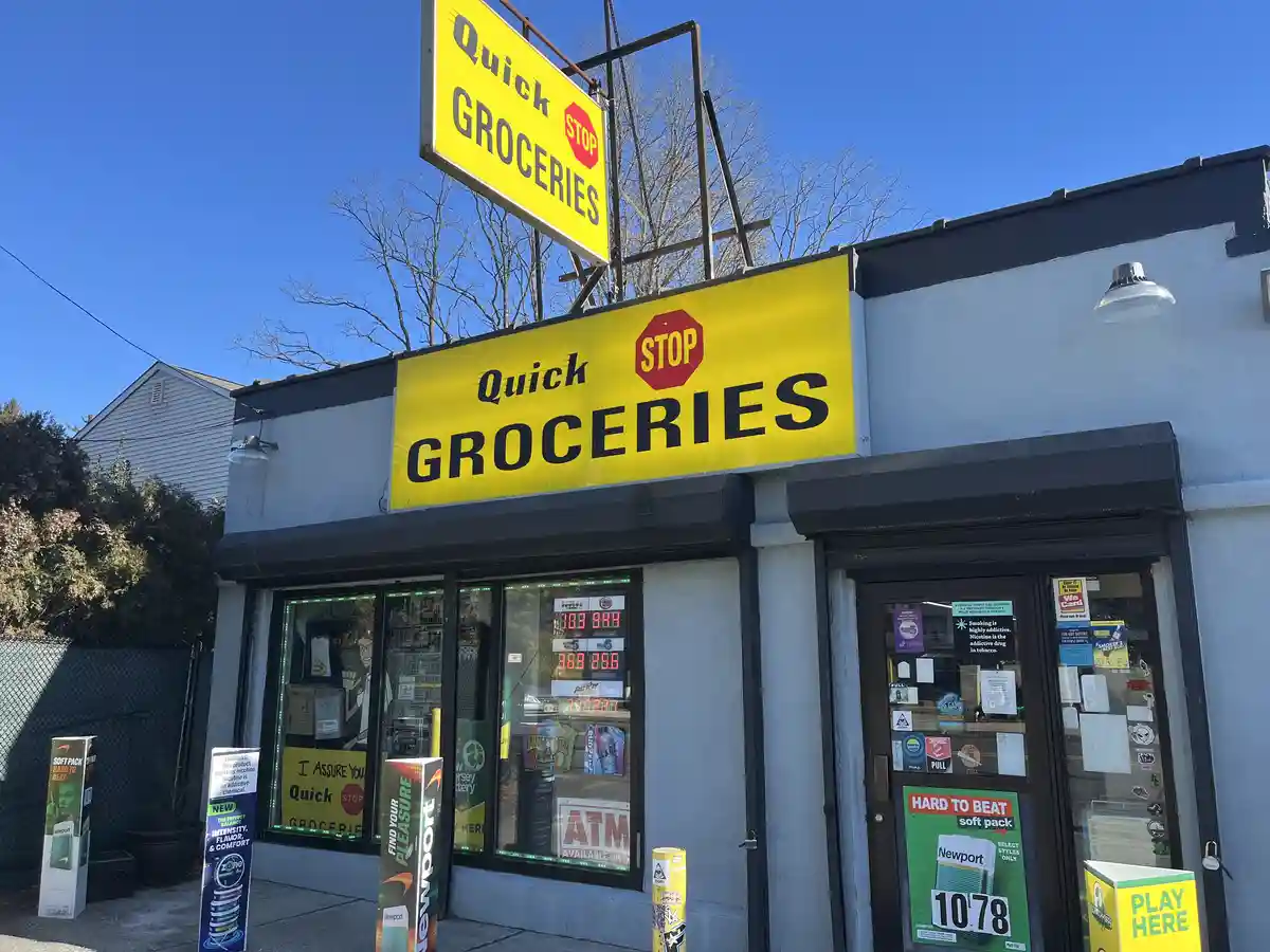 An exterior shot of a 'Quick Stop Groceries' store with a bright yellow sign featuring a red stop sign logo. The storefront…