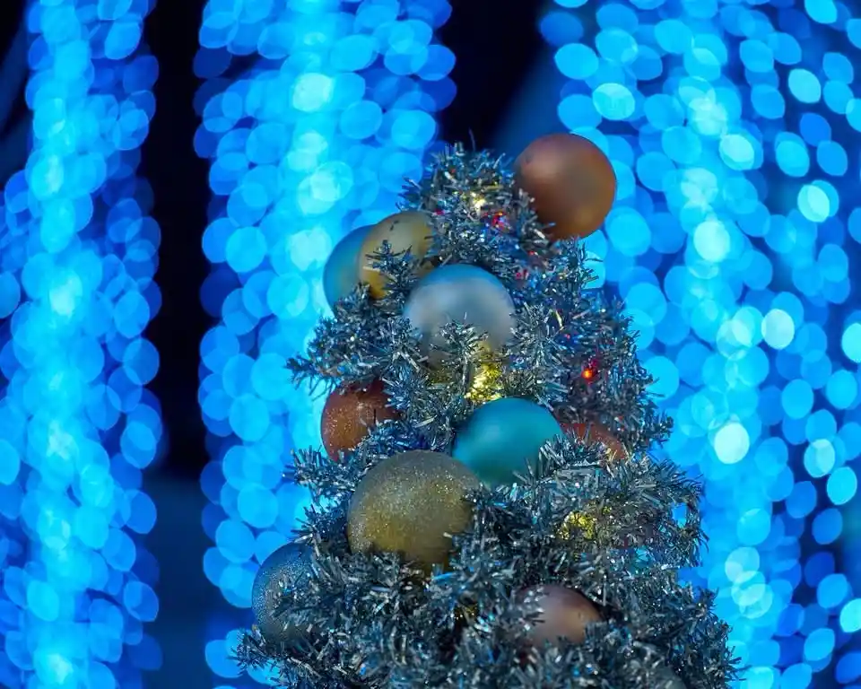 A small, decorated artificial tree with silver tinsel and multicolored ornaments stands against a blurred background of…