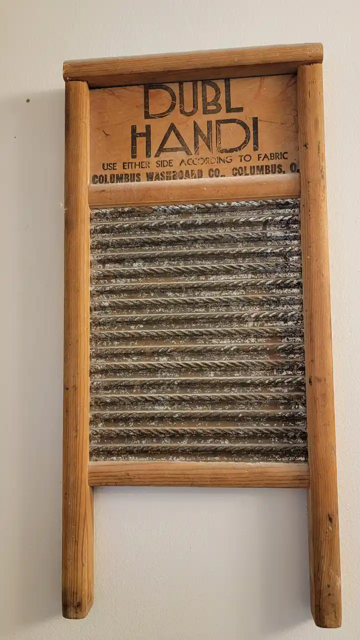 A vintage wooden washboard with a metal corrugated surface is mounted on a white wall. The top panel has faded black text…