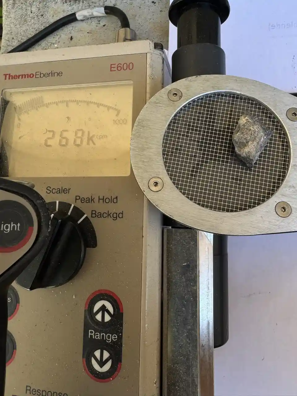 A close-up photo shows a Thermo Eberline E600 radiation detector with its analog display reading '258k cpm'. A rough…