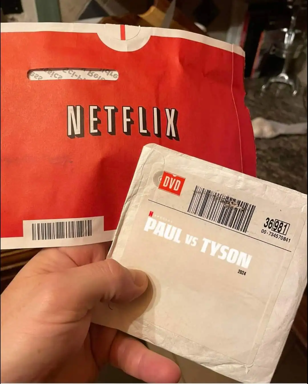A hand holds a white DVD case labeled 'PAUL VS TYSON 2024' in front of a red Netflix mailer bag, humorously contrasting…