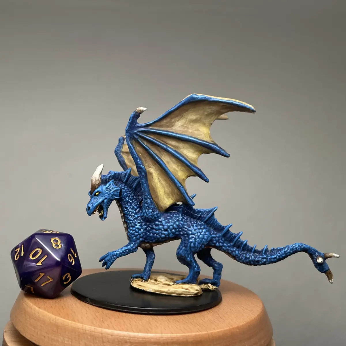 A blue dragon miniature with gold wings and horns stands on a black base, positioned next to a purple 20-sided die showing…