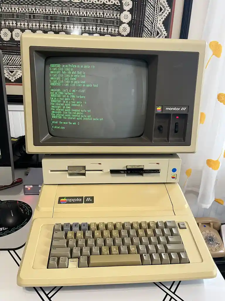 A vintage beige Apple IIe computer with a CRT monitor displays a green terminal chat log where users joke about not being…