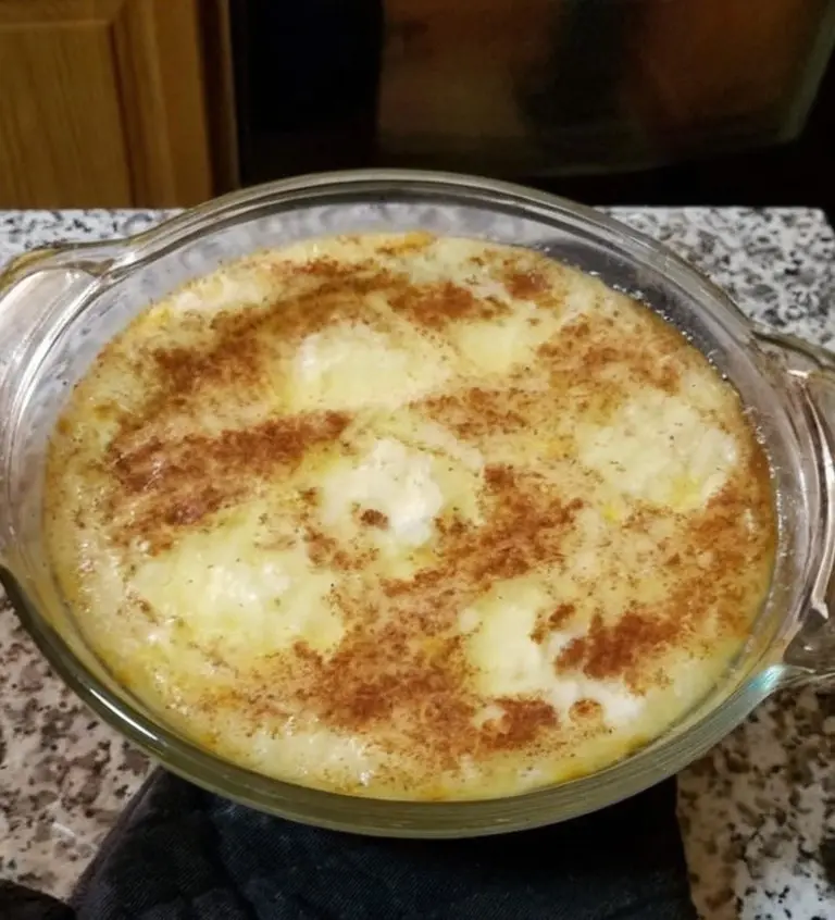 A close-up photo shows a round, glass baking dish containing a creamy, custard-like dessert topped with a sprinkle of…