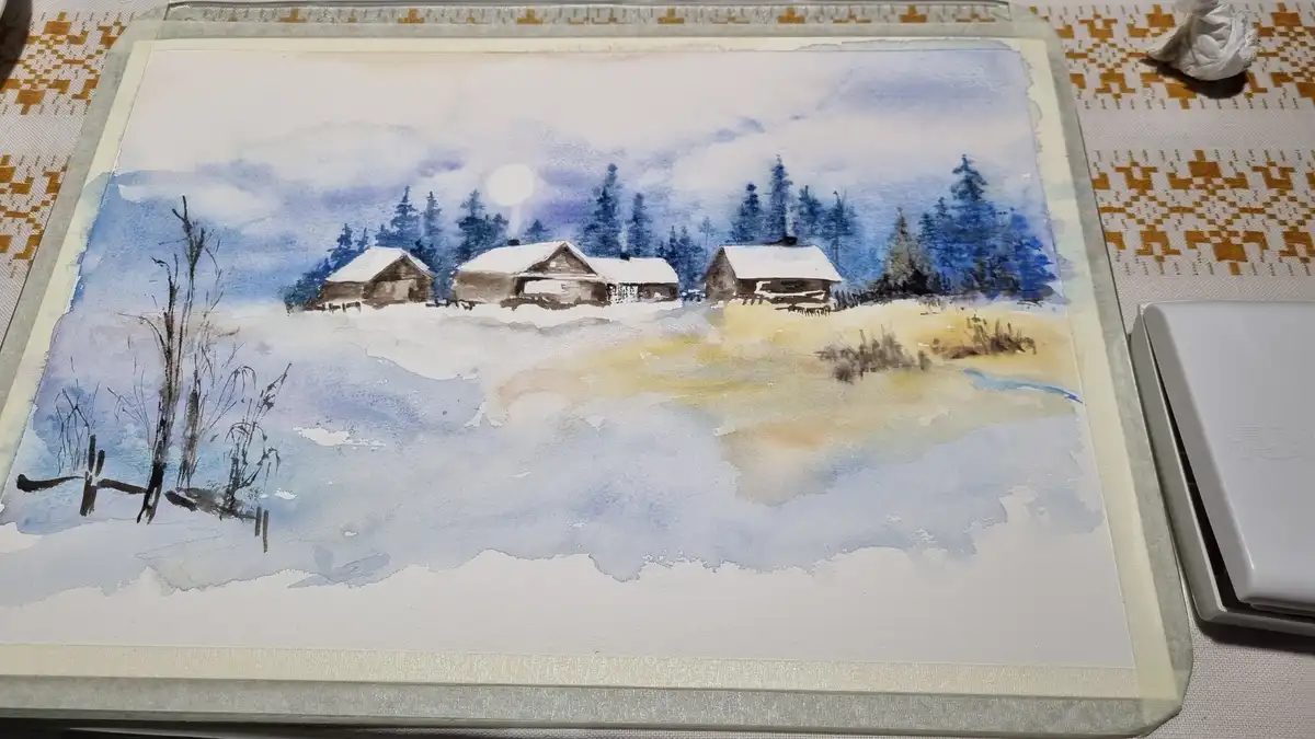 A watercolor painting of a snowy winter scene with three cabins, evergreen trees, and a full moon, displayed on a table next…