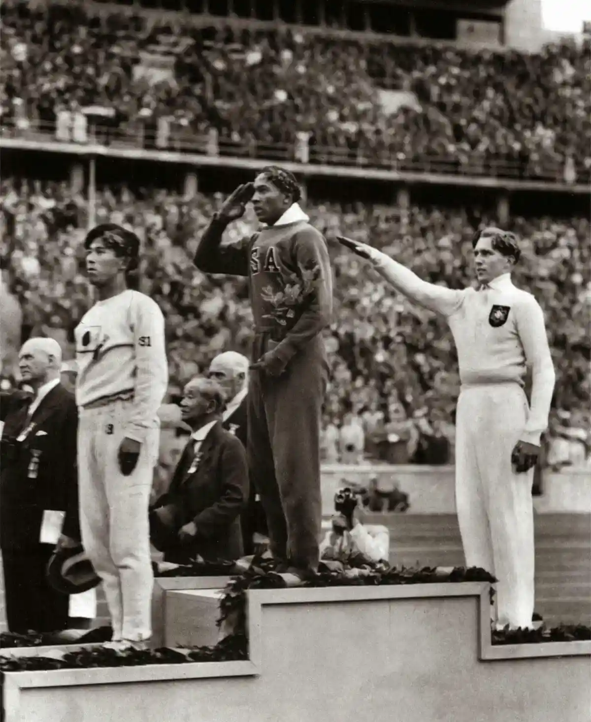 A black-and-white photo shows three male athletes standing on a podium at an Olympic ceremony, saluting. The central…