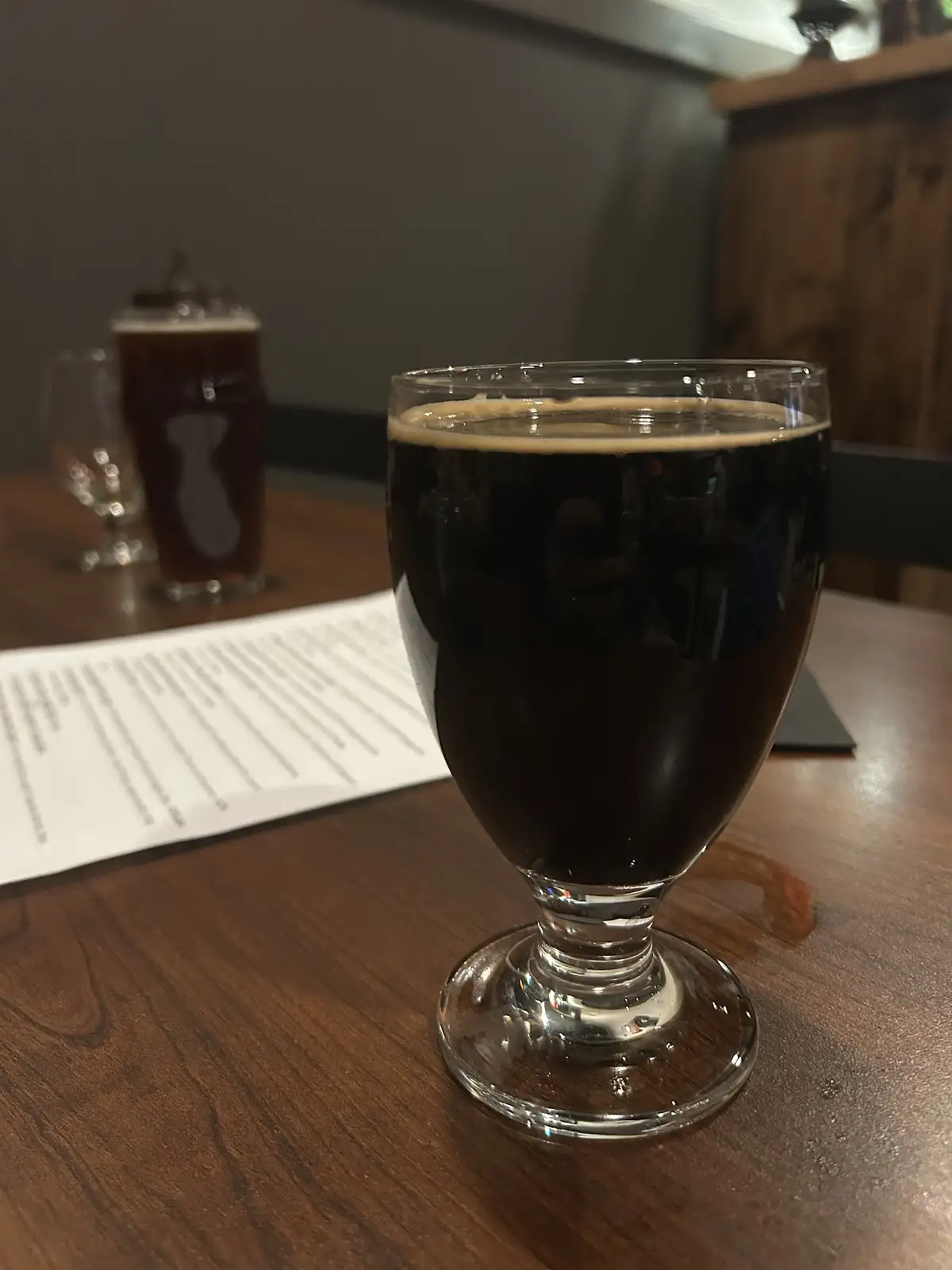 A close-up photo shows a dark, opaque beer in a stemmed glass on a wooden table, with a second beer and a menu blurred in…