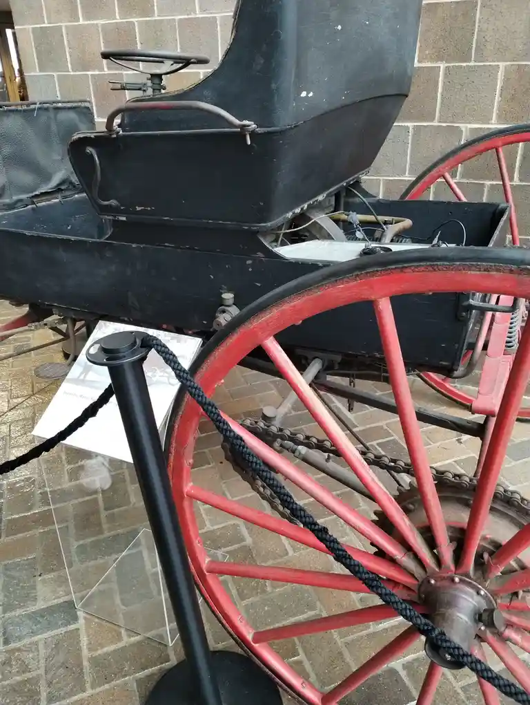 A close-up photo of an antique black horseless carriage with red spoked wheels, displayed in a museum behind a black rope…
