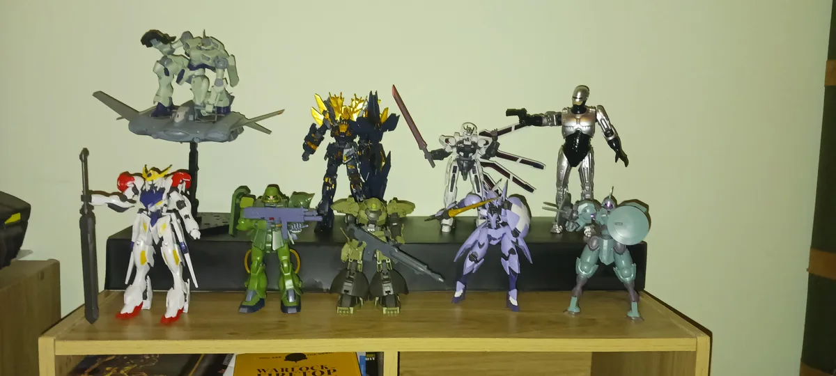 A collection of various robot action figures, including mechs and humanoid robots, are displayed on a wooden shelf against a…