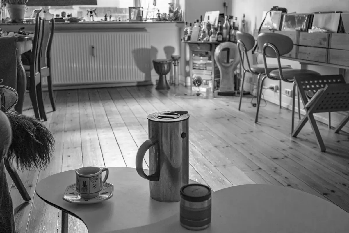 A black-and-white photo shows a small table with a coffee cup, a thermos, and a small jar. In the background, a room with…
