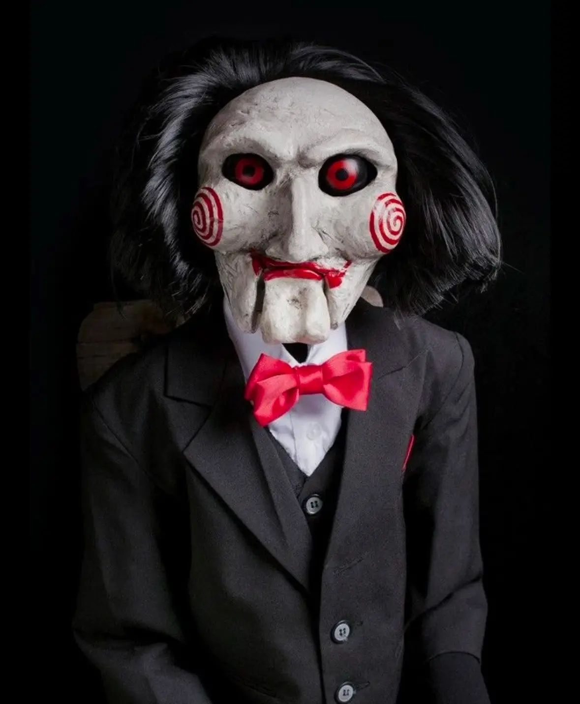 A person wearing a Jigsaw mask from the 'Saw' movie franchise, dressed in a formal black suit with a white shirt and a…
