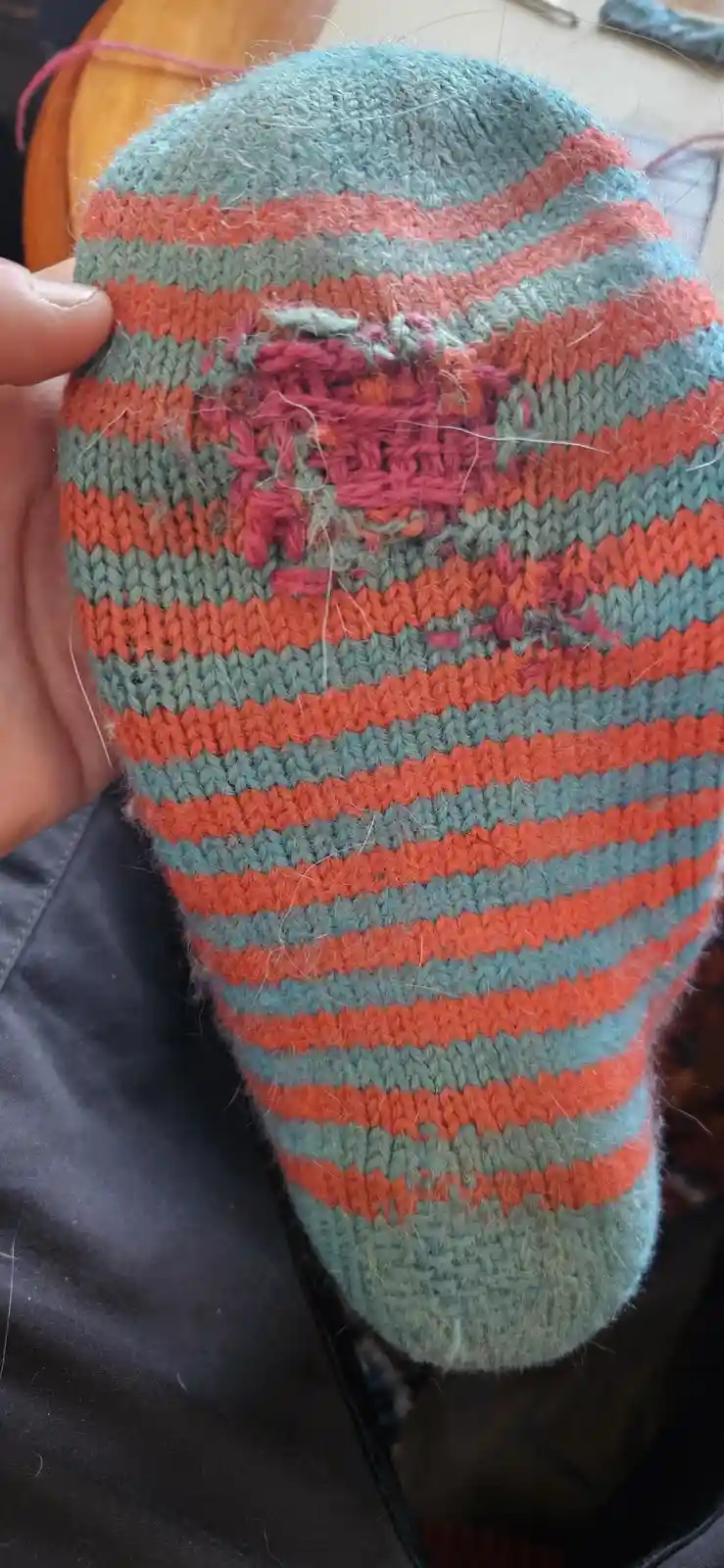 A hand holds a hand-knitted sock with alternating orange and teal stripes, showing a large, messy hole in the fabric where…