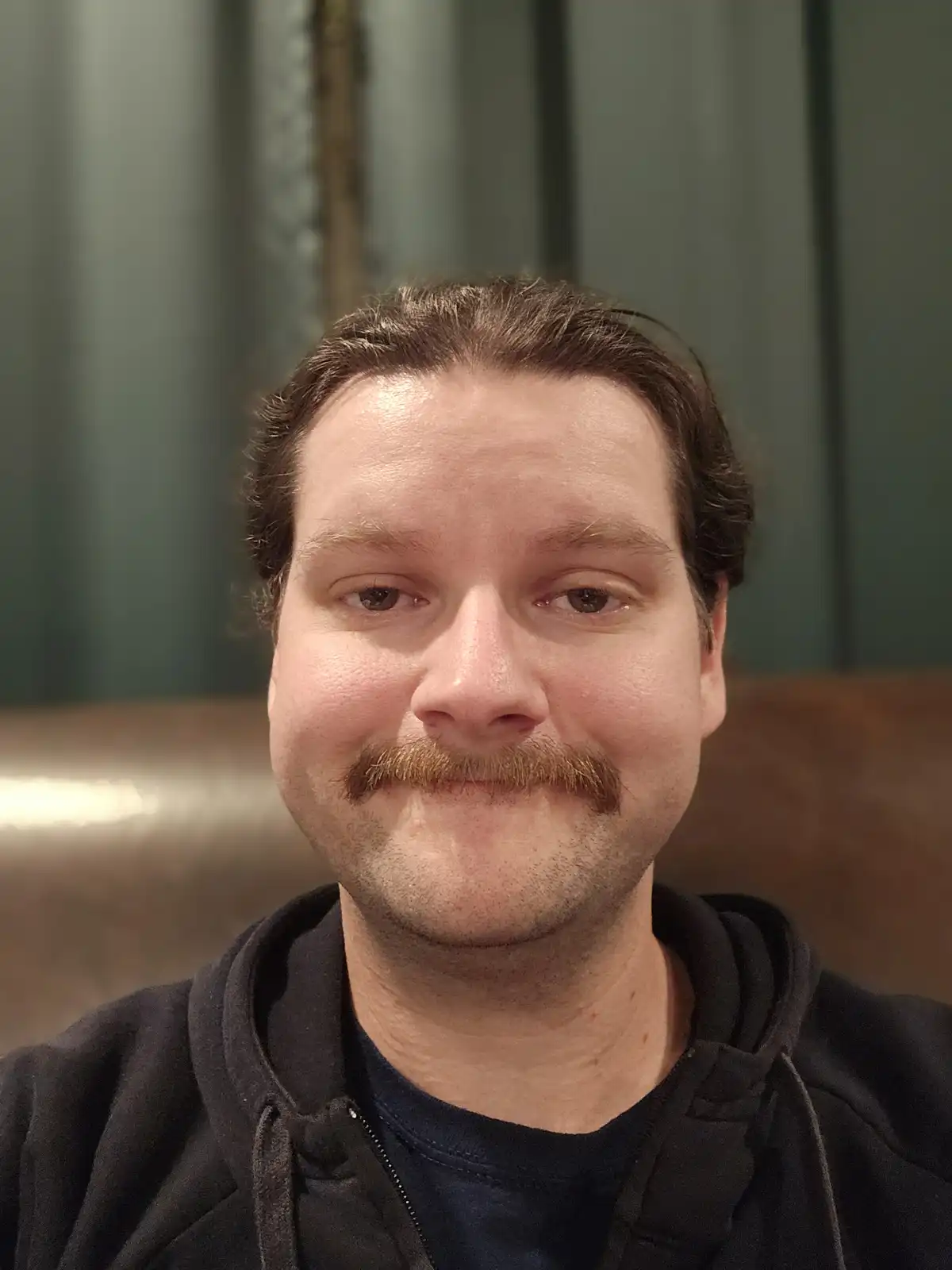 A close-up selfie of a man with a thick, reddish-brown handlebar mustache, wearing a black hoodie over a dark t-shirt…