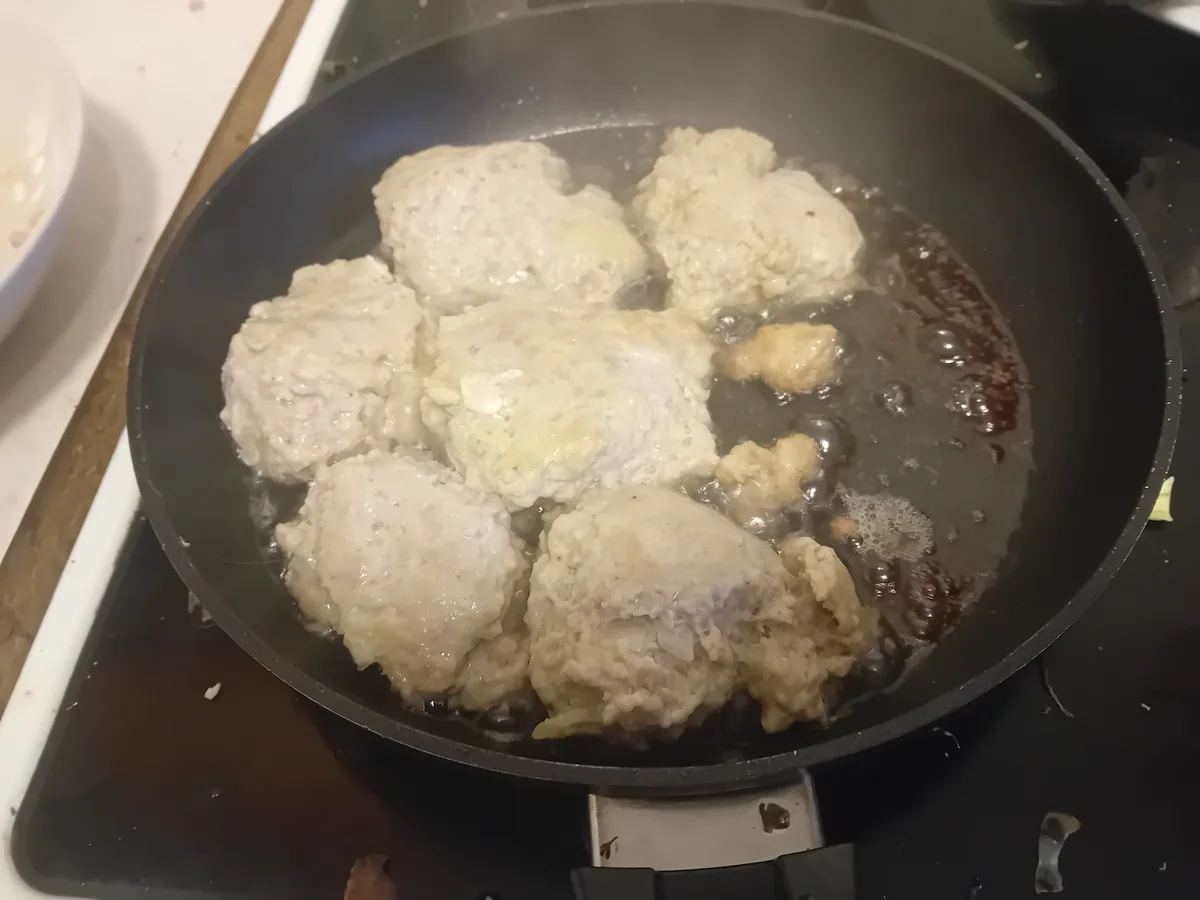 A black frying pan on a stovetop contains several lumpy, pale, uncooked-looking meatballs sizzling in oil. The meatballs are…