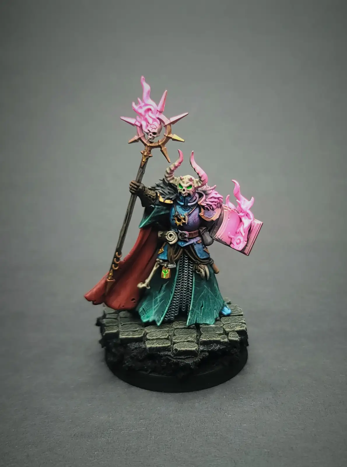 A painted miniature of a demonic-looking wizard with glowing green eyes, pink horns, and a skull face, holding a staff…