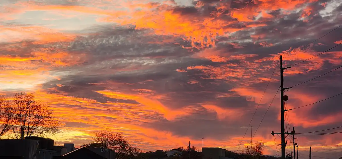 A vibrant, fiery orange and red sunset fills the sky above a suburban landscape with silhouetted trees, buildings, and a…