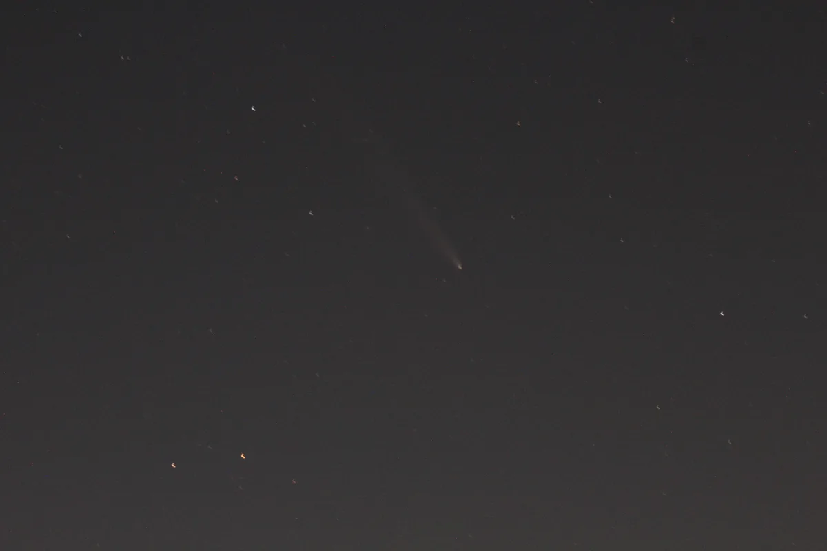 A dark night sky filled with faint stars and light pollution, featuring a single, blurry comet with a visible tail streaking…