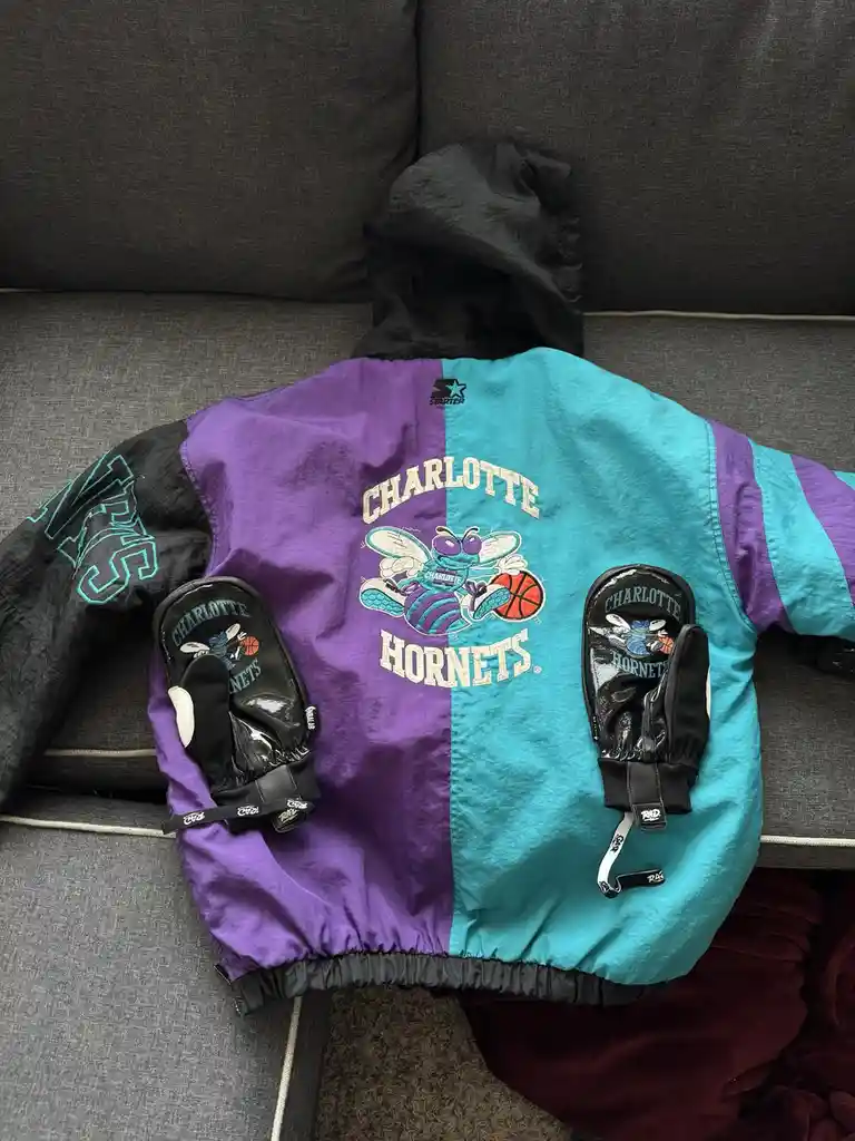A vintage Charlotte Hornets Starter jacket, split purple and teal, lies on a gray couch with two matching black gloves…