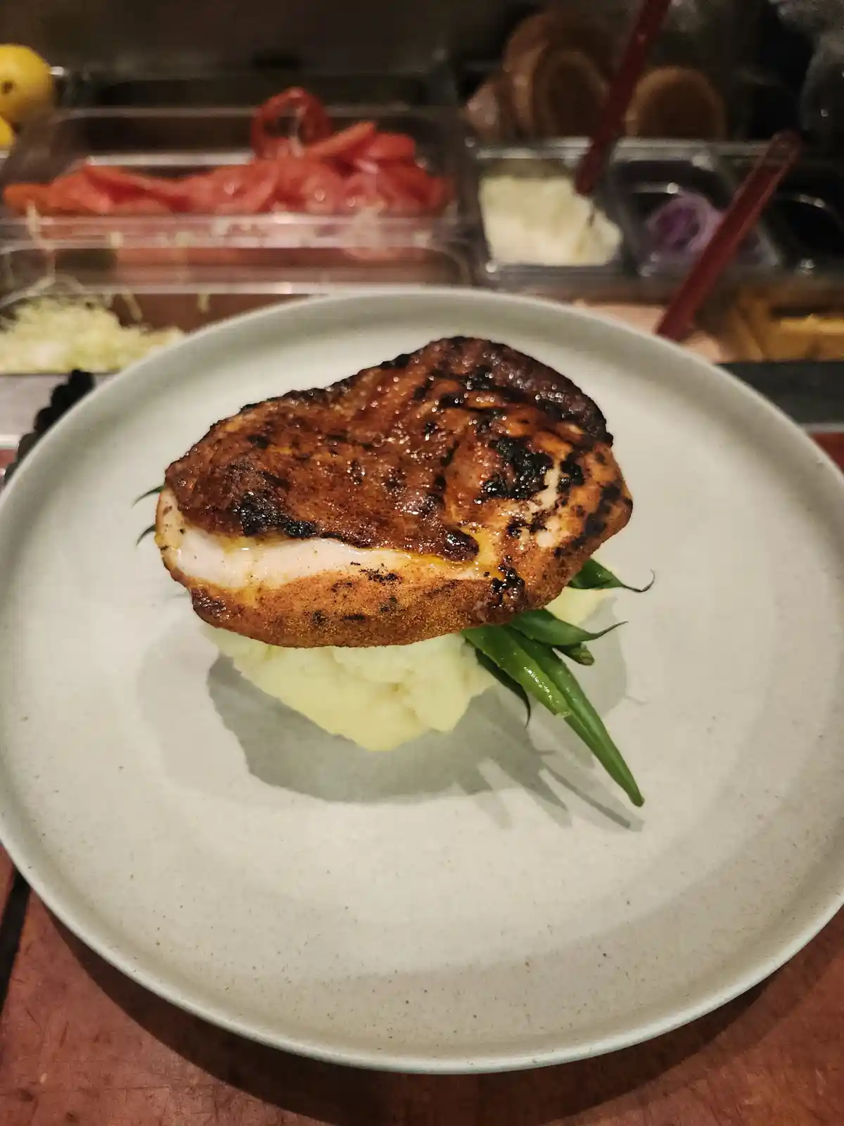 A close-up photo of a grilled chicken breast served on a bed of mashed potatoes and green beans on a light gray plate. In…