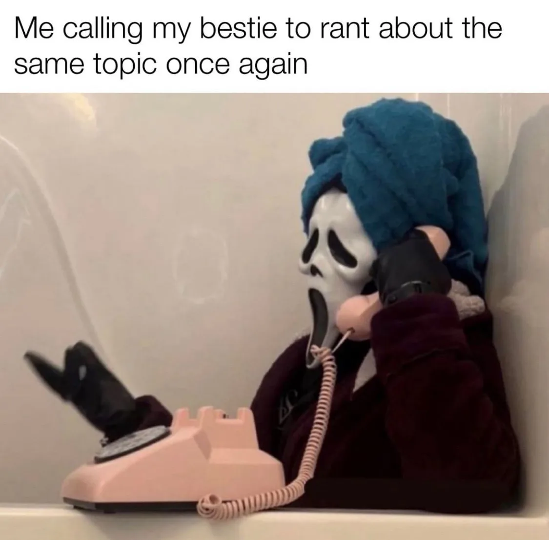 A person wearing a white Ghostface mask from 'Scream', a blue towel on their head, and a dark robe is sitting in a bathtub…