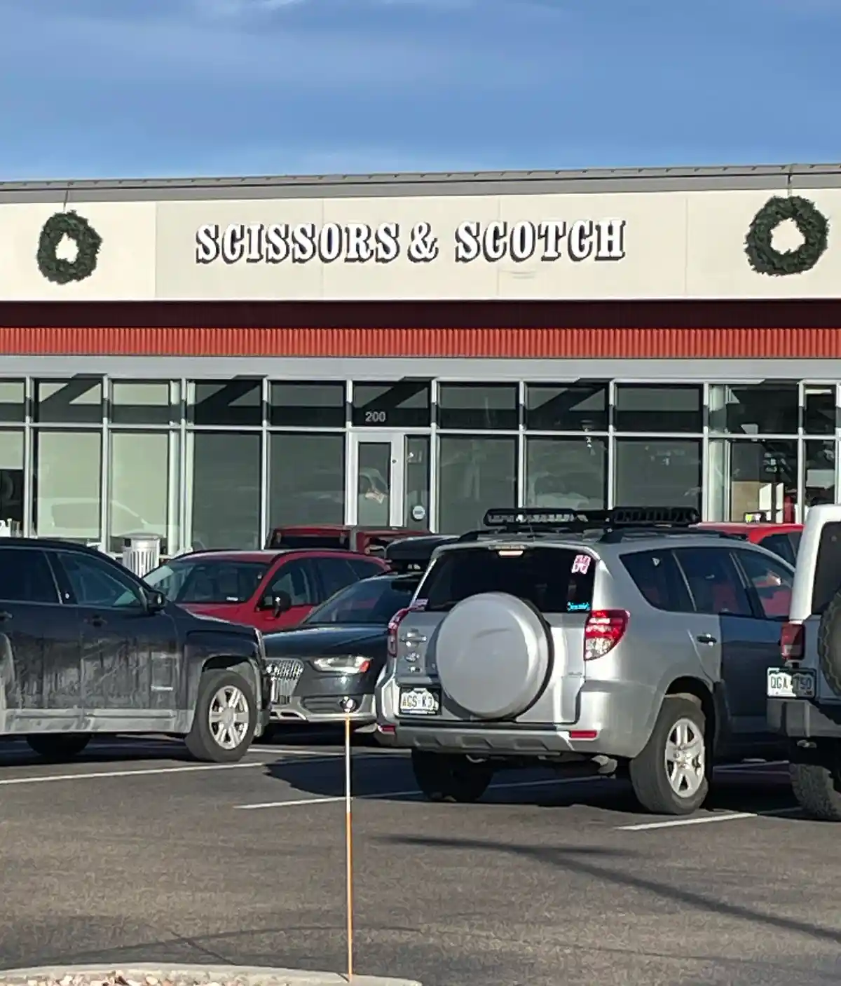 An exterior shot of a storefront with a sign reading 'SCISSORS & SCOTCH' flanked by two green wreaths. Several cars are…