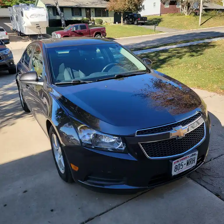 A black Chevrolet Cruze sedan is parked on a residential driveway, its glossy hood reflecting trees. The Wisconsin license…