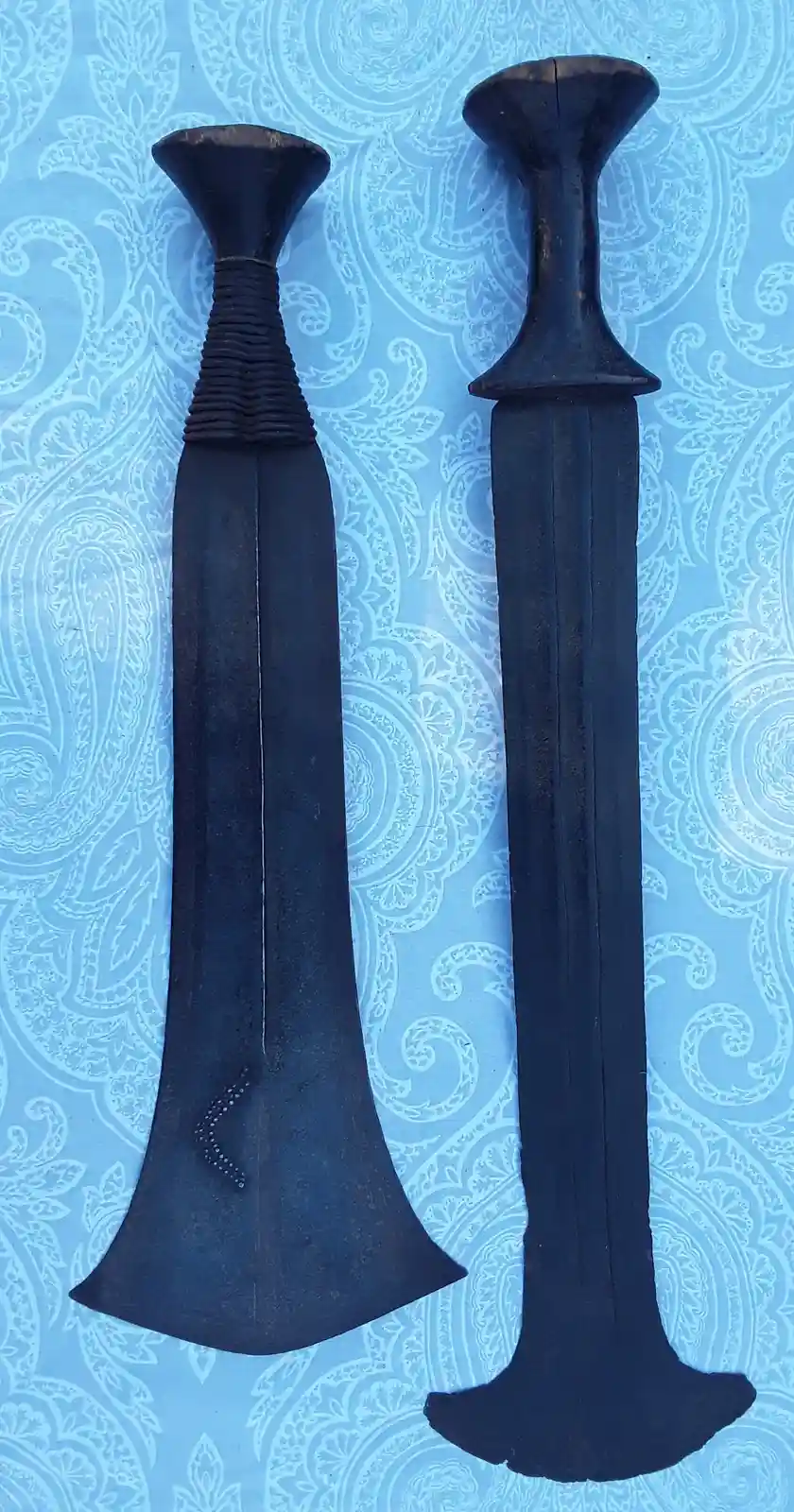 Two black kris blades with ornate, flared hilts and decorative patterns are displayed vertically side-by-side against a blue…