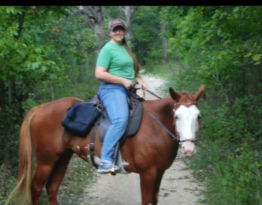 A woman in a green shirt and blue jeans is smiling while riding a brown horse with a white face along a dirt trail through a…
