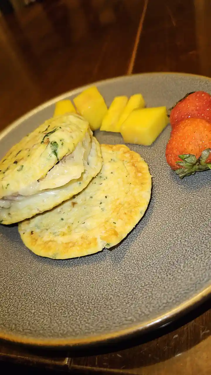A plate of food featuring a folded omelet with a visible filling, accompanied by diced yellow fruit and two whole…