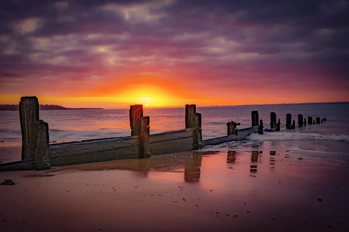 A serene beach scene at sunset with vibrant orange and purple skies reflected in wet sand, featuring a decaying wooden pier…