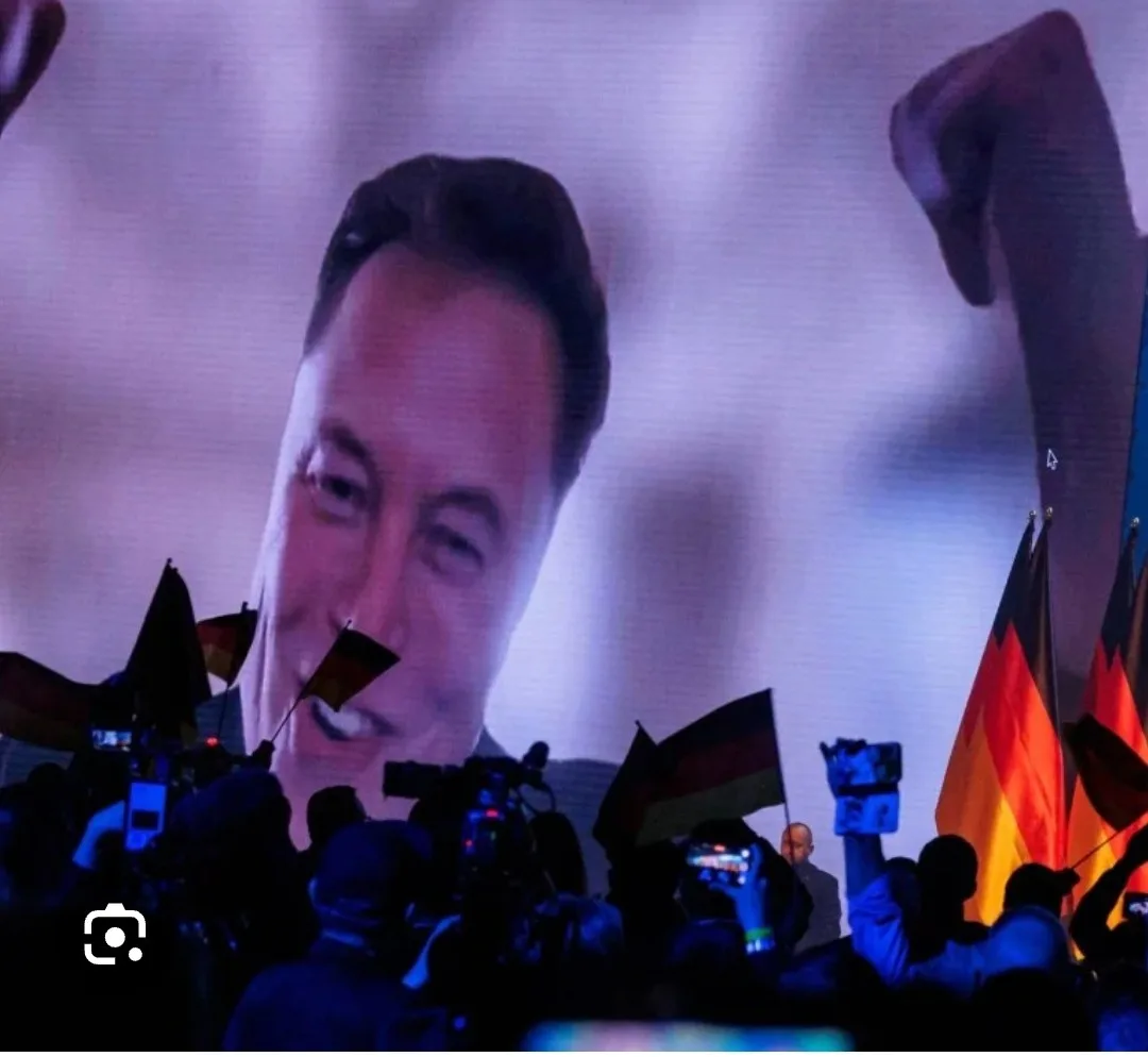 A large screen displays a smiling Elon Musk with his fist raised, while below, a crowd holds German flags and phones, with a…