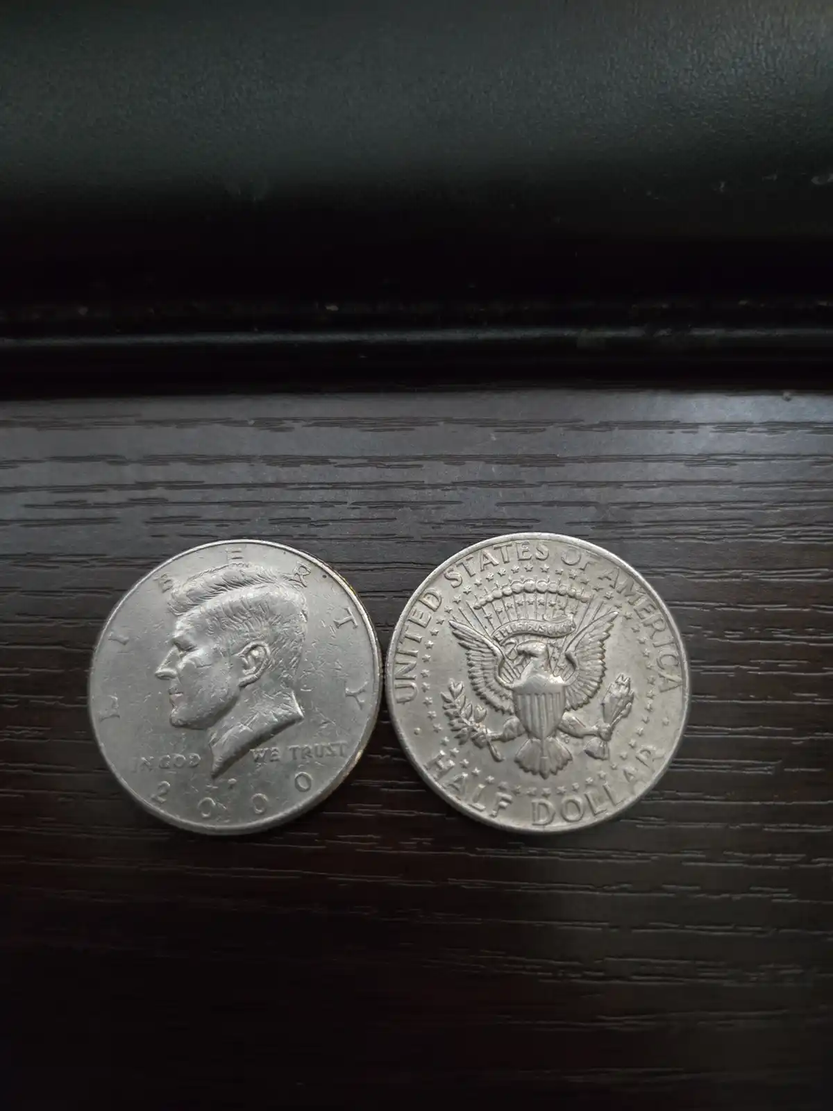 Two silver U.S. half dollars are placed side by side on a dark wooden surface; the left coin shows a profile of John F…