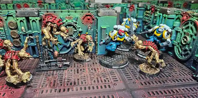 A photo of painted miniature wargaming figures: several grotesque, biomechanical alien creatures in red and gold armor clash…