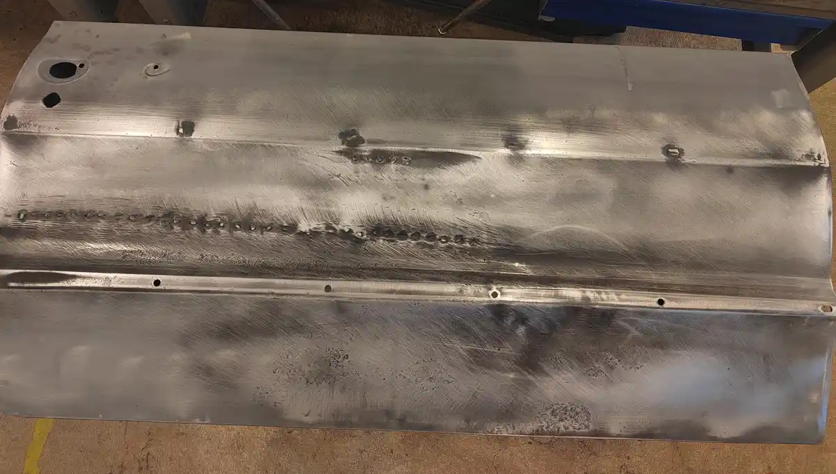 A close-up photo of a brushed metal automotive panel with visible weld beads and holes, resting on a workshop floor. The…