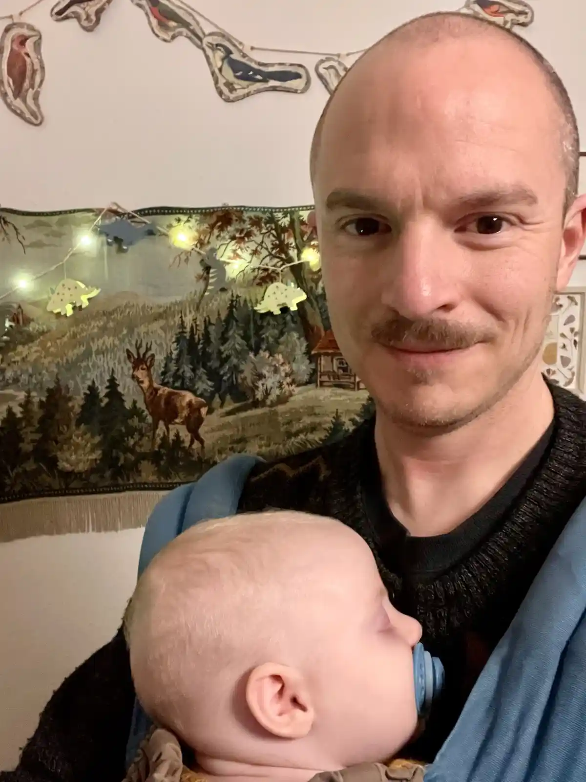 A man with a mustache and shaved head smiles at the camera while holding a sleeping baby in a blue carrier. The baby has a…