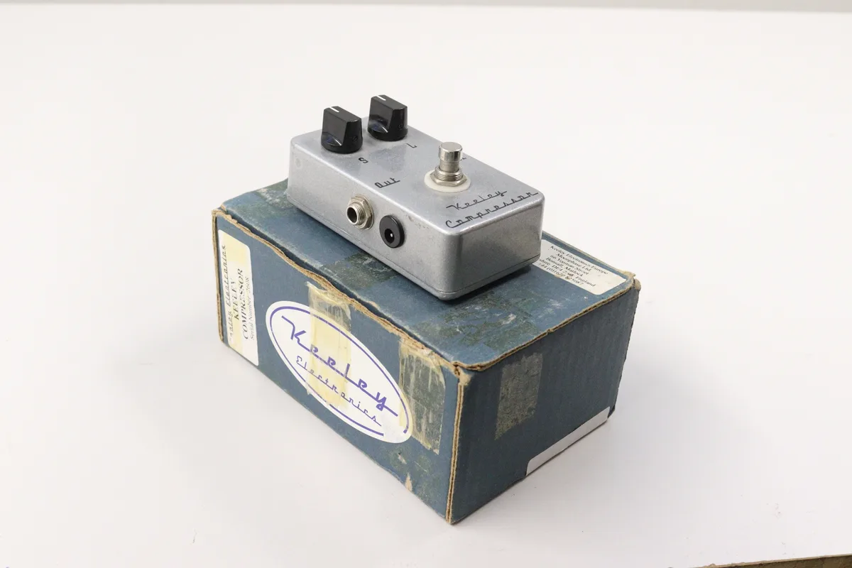 A silver Keeley Compressor guitar effects pedal with two black knobs and a footswitch sits atop its original blue cardboard…