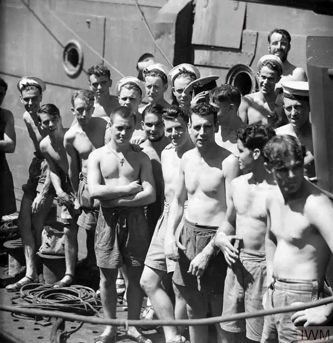 A black-and-white historical photograph shows a group of shirtless young men, likely sailors, posing on the deck of a ship…