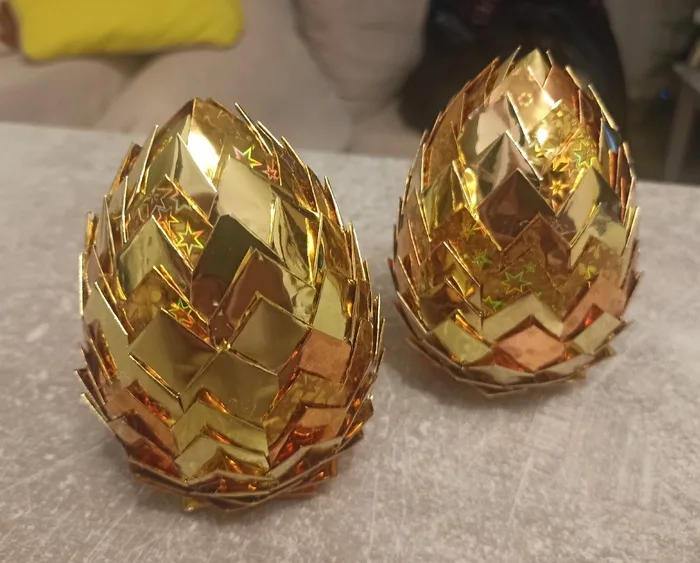Two ornate, golden, spiky eggs made of reflective foil sit side by side on a gray surface. The eggs have a geometric…