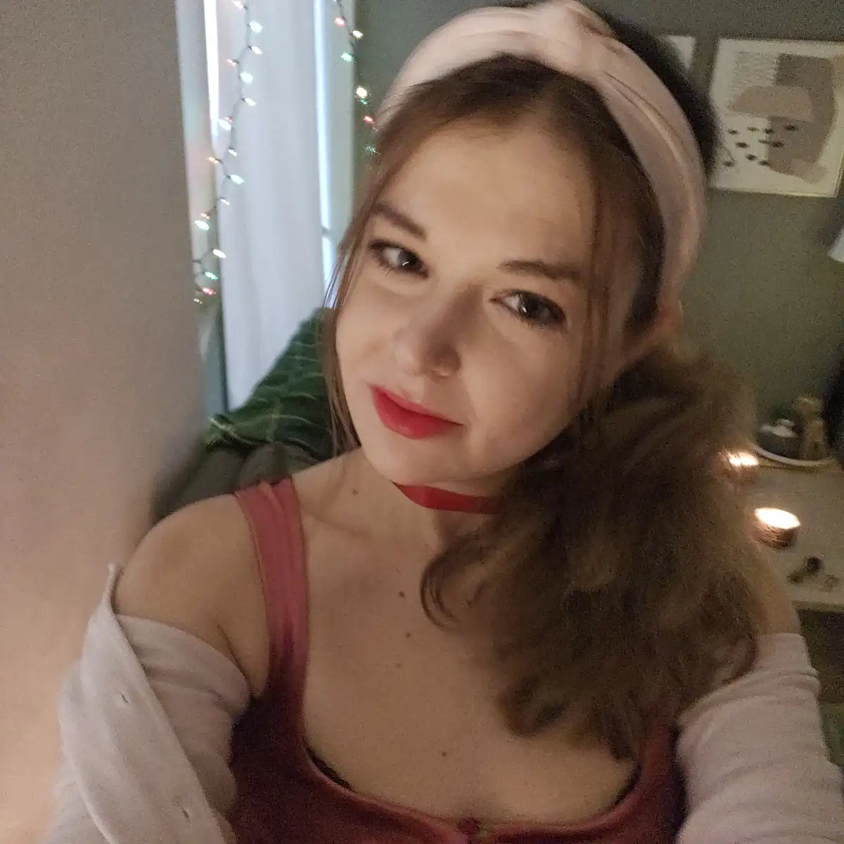 A young woman with brown hair wearing a white headband, red lipstick, and a red top under a white shirt, looks at the camera…