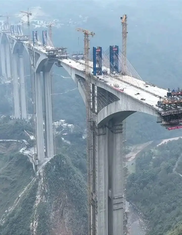aerial view of a massive, partially constructed concrete bridge spanning a deep valley between mountains, with cranes and…
