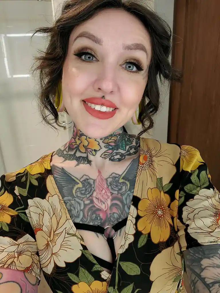 A woman with extensive tattoos covering her neck and chest, wearing a floral-patterned top and bright yellow hoop earrings…