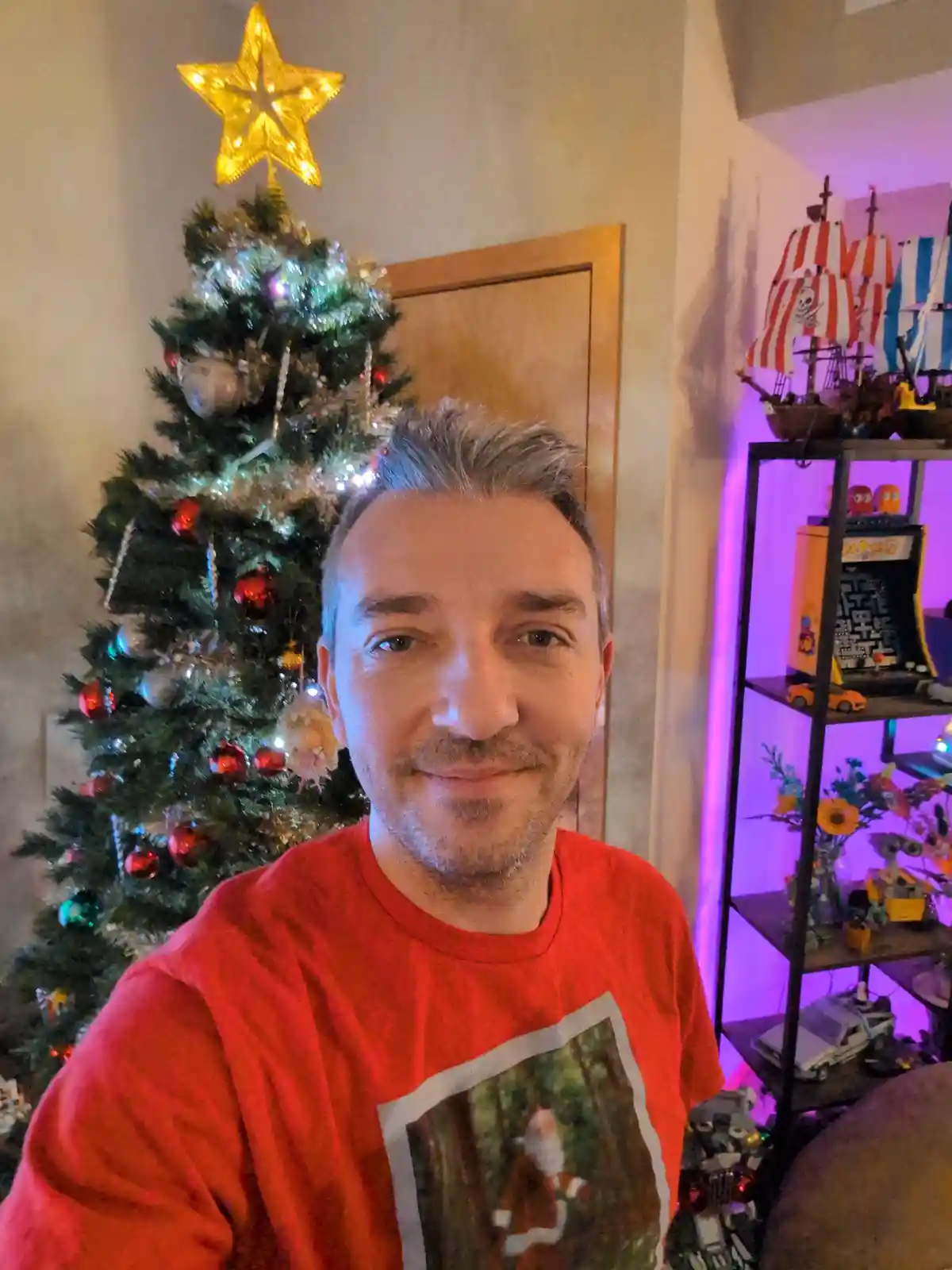 A man with gray hair and a red t-shirt featuring a Santa Claus image smiles at the camera while standing in front of a…