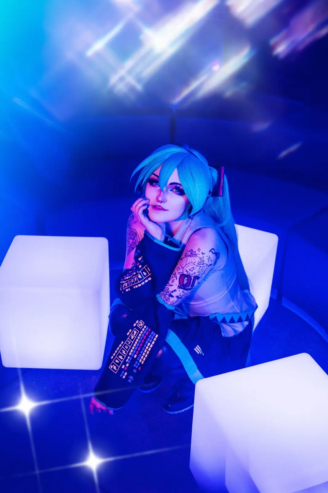 A person in a cosplay of Hatsune Miku sits on a white cube in a blue-lit room with disco lights, wearing a sleeveless top…