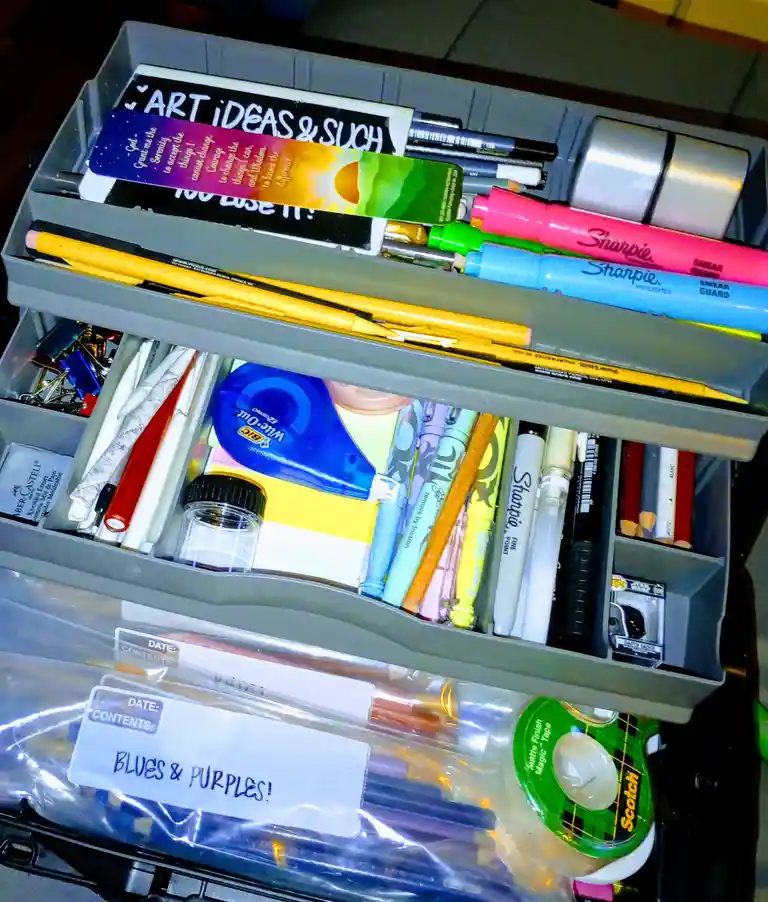 A grey multi-compartment organizer drawer filled with art supplies like Sharpie markers, pencils, tape, and pens. A label on…
