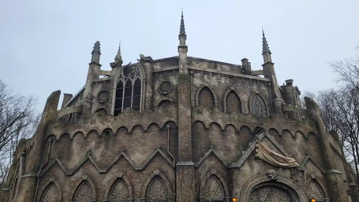 A low-angle shot of a dilapidated, gothic-style brick building with broken windows, crumbling stonework, and pointed spires…