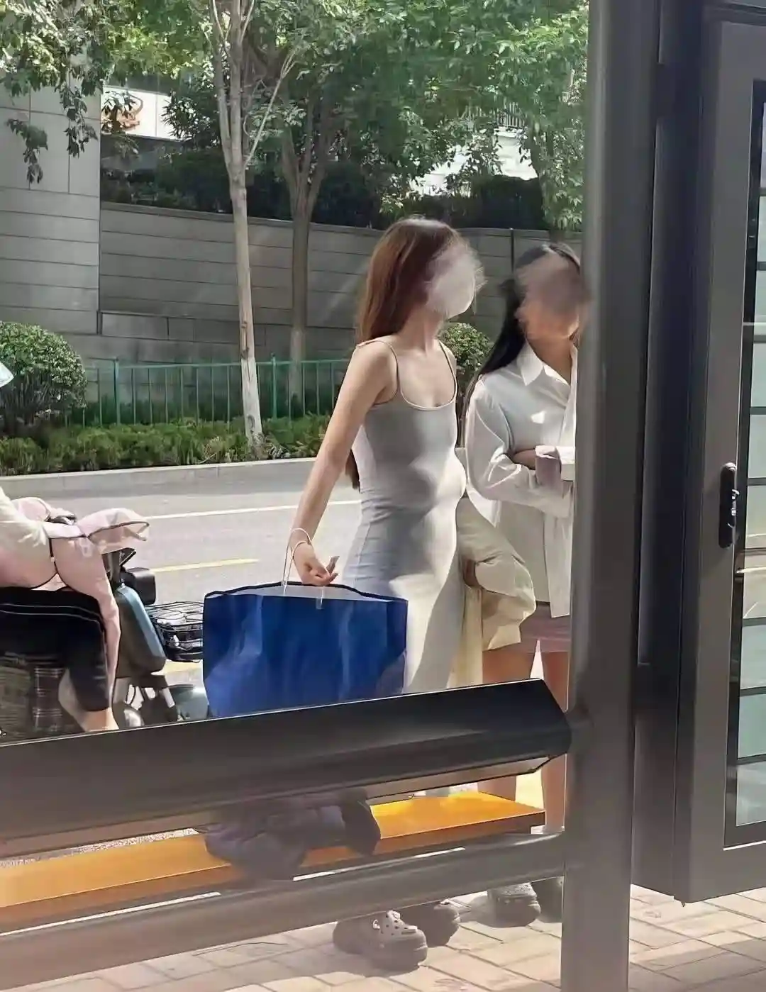 Two women stand outside a building entrance, one in a form-fitting grey dress holding a blue shopping bag, the other in a…