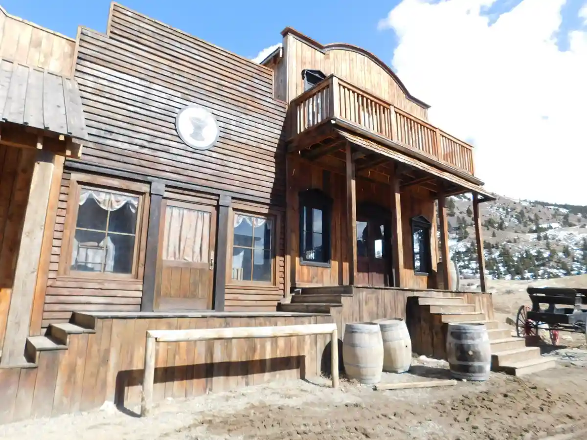 a wooden western-style saloon with a porch, barrels, and a martini glass logo on the wall, set against a snowy mountain…